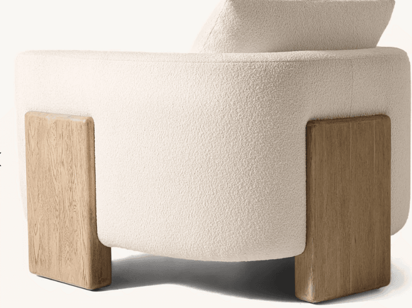 A close-up, side-view of an armchair featuring a plush, textured boucle upholstery in an off-white or cream color. The armchair has a rounded, organic form. It is supported by two distinct, chunky wooden legs or base elements that extend slightly beyond the upholstered body, creating a visual contrast in materials and form. The wood appears to be a light to medium tone with visible grain. — 3D model