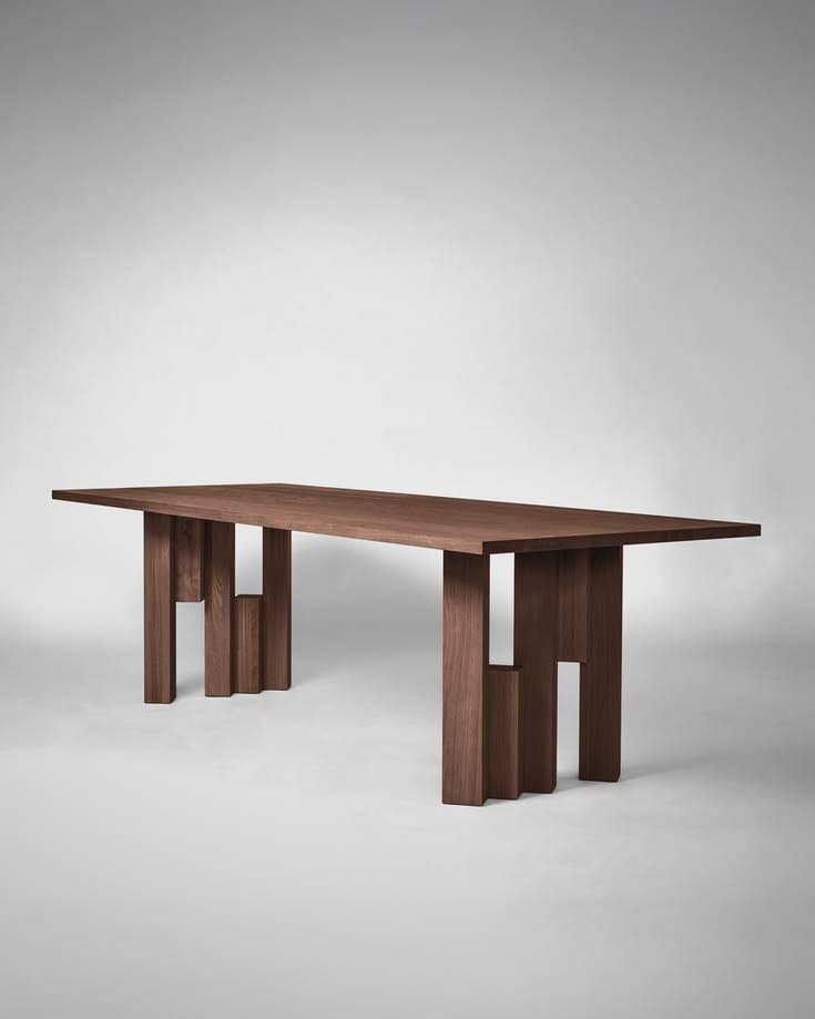 A modernist dining table crafted from dark brown walnut wood. The table features a substantial rectangular top with clean edges, supported by two unique, sculptural bases. Each base is composed of multiple solid rectangular blocks of wood, arranged in an interlocking, geometric pattern that creates a visually striking and stable support structure. The overall aesthetic is minimalist yet impactful, highlighting the natural beauty and grain of the wood through its simple, strong forms. — 3D model