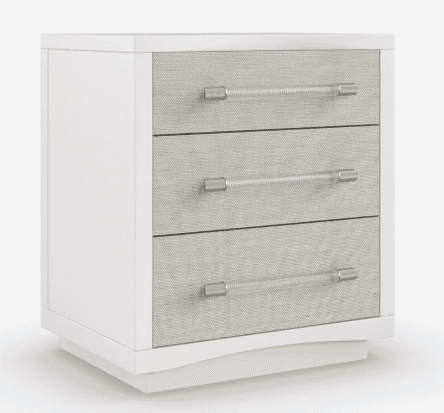 A contemporary white nightstand featuring three drawers with textured, light-colored fabric fronts and clear acrylic bar pulls with polished chrome accents. The cabinet sits on a subtle, recessed plinth base. — 3D model