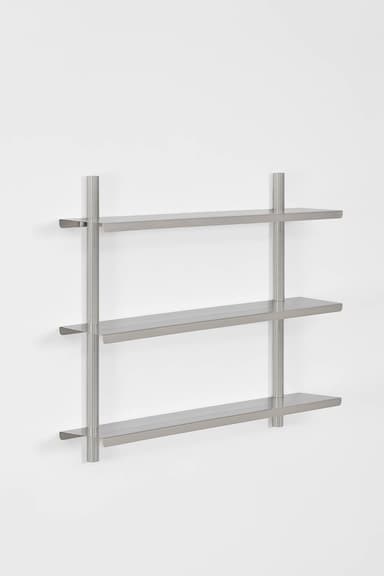 A modern, minimalist wall-mounted shelf unit featuring three rectangular shelves made of brushed silver-gray metal. The shelves are supported by two vertical cylindrical rods that extend from the floor to above the top shelf, where they feature keyhole-style wall mounting brackets. The design is sleek and industrial. — 3D model
