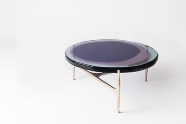 A round coffee table featuring a clear glass top with an iridescent finish that displays shifting colors, predominantly purple and blue tones. Beneath the glass is a thin black rim. The table is supported by a sculptural three-legged metal base with a warm, brass-like finish. The legs taper downwards and meet in a central point, giving the table a distinctive, modern appearance. — 3D model