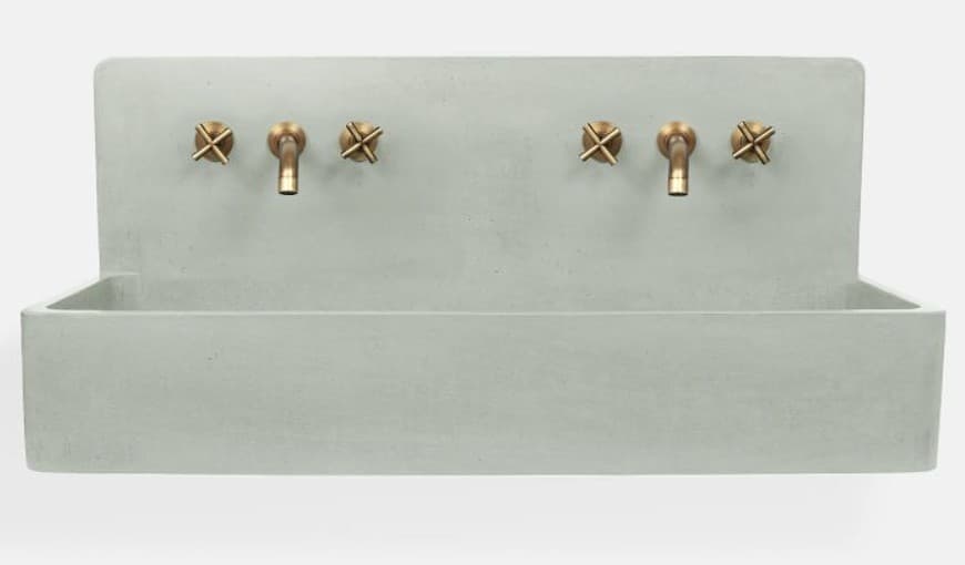 A minimalist, double basin, wall-mounted sink crafted from light grey concrete, featuring two sets of wall-mounted brass faucets. Each faucet set consists of a central spigot and two 'X'-shaped knob handles, all in a warm, patinated brass finish. The sink's form is a rectangular trough with a high back panel where the plumbing fixtures are integrated, presenting a clean, industrial aesthetic. The concrete surface appears smooth and uniform, indicative of modern casting techniques. — 3D model