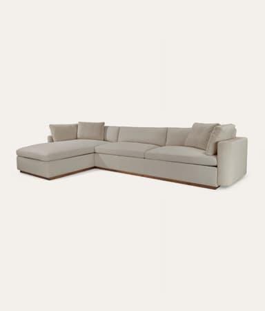 An L-shaped sectional sofa featuring a chaise lounge on one end and a multi-seat configuration. The sofa is upholstered in a light gray or beige fabric and sits on a recessed, dark wooden plinth base. It has a modern, minimalist design with clean lines and includes several loose back cushions and accent pillows. — 3D model