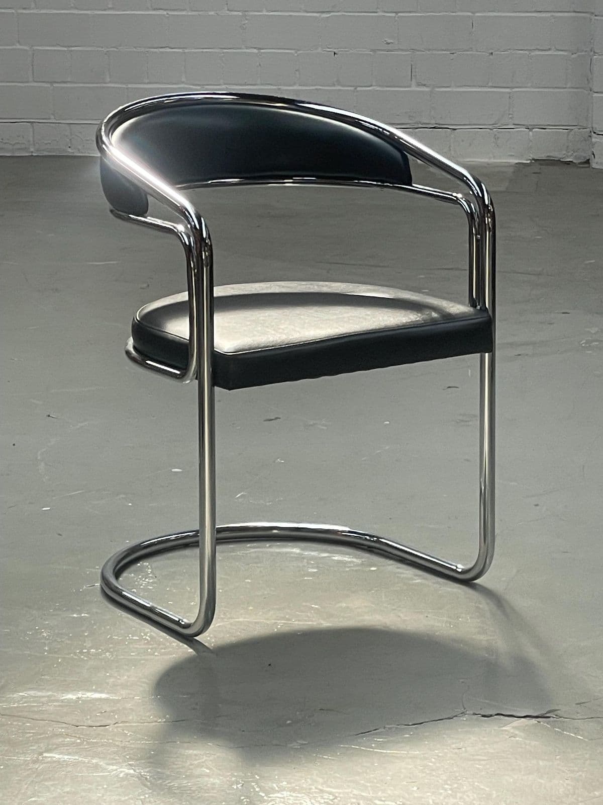 A modern armchair with a tubular metal frame and upholstered seat and backrest. The frame has a chrome finish and features a distinctive cantilevered base. The seat and backrest are padded and covered in black material, likely faux leather or vinyl. The armrests are integrated into the frame. — 3D model