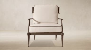 A contemporary lounge armchair featuring a dark-toned exposed metal frame and plush, light-colored upholstered cushions. The chair has a distinctive backrest with a wide, curved top rail and vertical support elements, complemented by straight, padded armrests. The legs are subtly tapered, contributing to a sleek and elegant profile. — 3D model