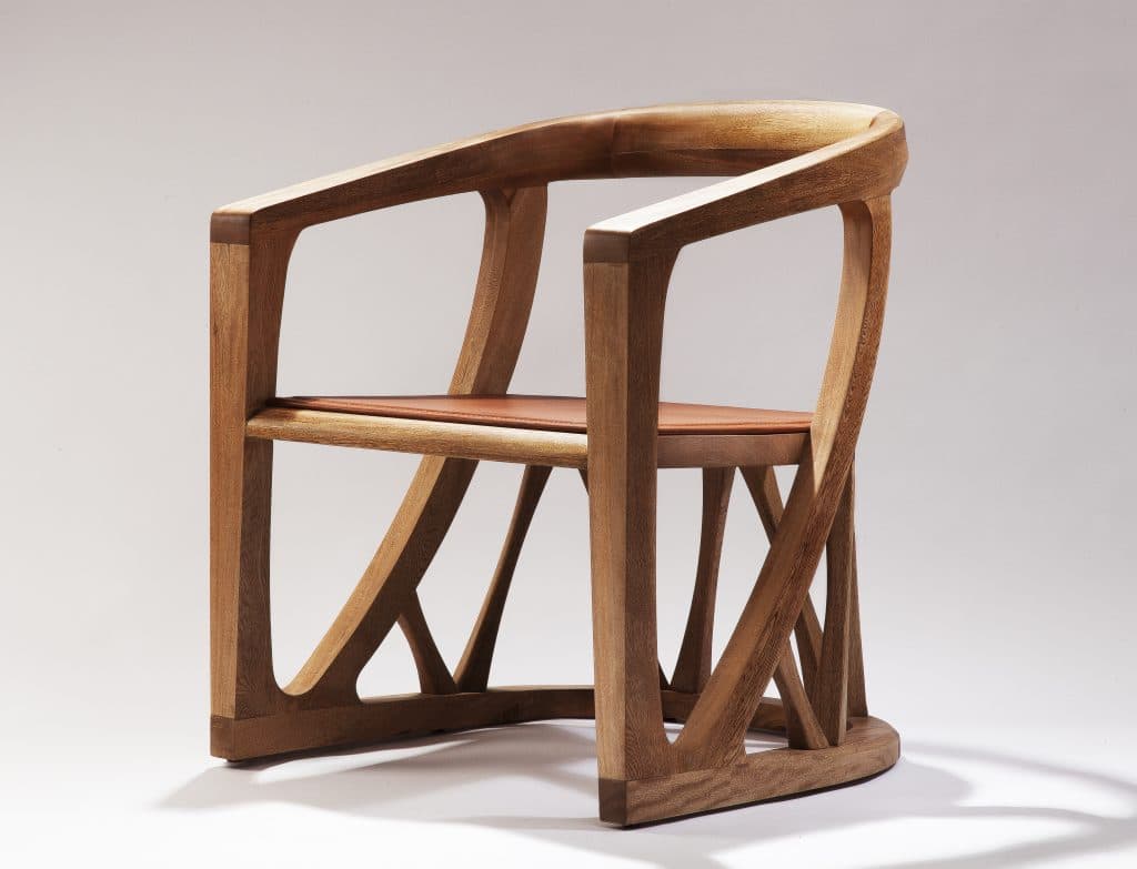 A contemporary armchair crafted from light-colored solid wood, featuring a gracefully curved backrest and integrated armrests. The chair incorporates a geometric, sculptural base with multiple intersecting wooden struts providing support. A contrasting reddish-brown leather cushion forms the seat, complementing the natural wood tone. — 3D model