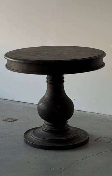 A small, round accent table featuring a dark, distressed finish and a single turned pedestal base. The tabletop is circular with a subtle rim, and the base consists of a baluster-like turned support leading to a broad, round foot. The overall aesthetic is rustic or traditional. — 3D model