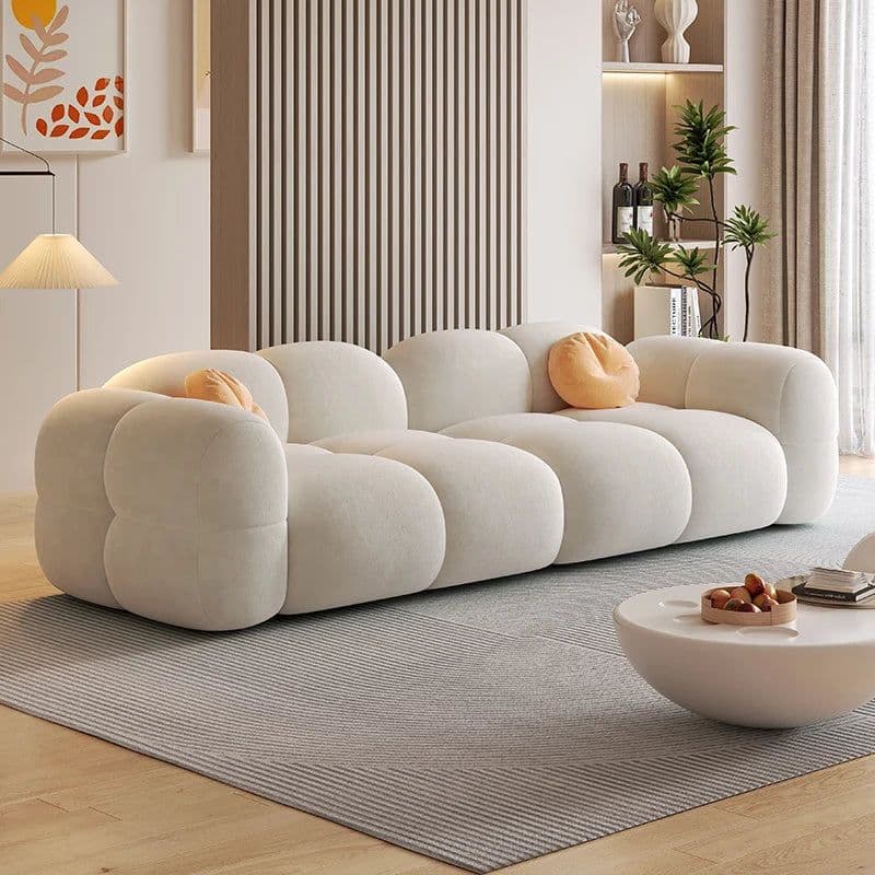A long, contemporary, off-white modular sofa featuring a distinctive bubbly design with rounded, bulbous cushions. The sofa has a low profile and a soft, inviting appearance, made from textured fabric. Two small, round, orange accent pillows are placed on the sofa. — 3D model