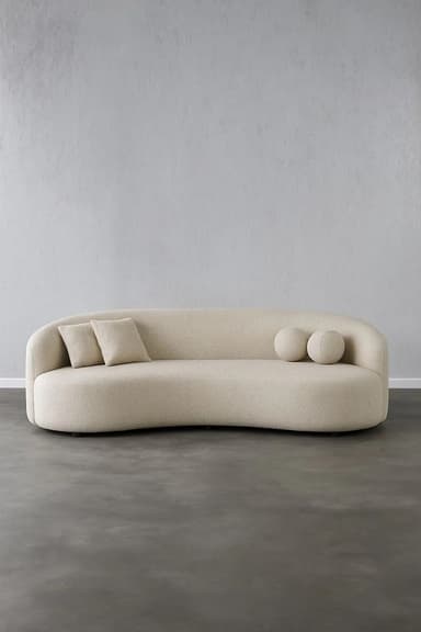 An asymmetrical curved sofa upholstered in a light beige, subtly textured fabric. It features a continuous, low-profile backrest and armrest, flowing into a single, organic form. The sofa is accompanied by two small rectangular throw pillows and two spherical decorative cushions, all in matching upholstery. — 3D model