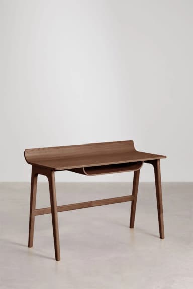 A minimalist wooden desk featuring a distinctive curved back panel and an integrated storage slot beneath the tabletop. The desk stands on four tapered, angled legs connected by a horizontal stretcher. — 3D model