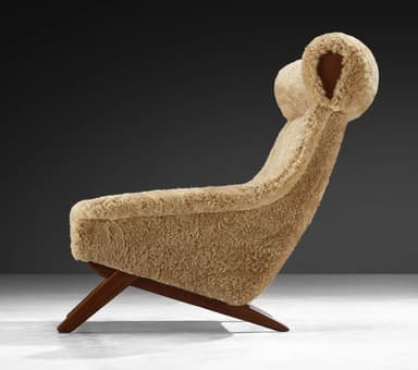 A high-backed armchair with a distinctive sloped silhouette. The chair is upholstered entirely in a textured, light brown sheepskin or shearling material, giving it a plush and organic appearance. The seat and back cushions are integrated into the overall form. The base consists of dark wood legs with a visible grain, angled outwards for stability. The headrest area is pronounced and rolls over the top of the backrest. — 3D model