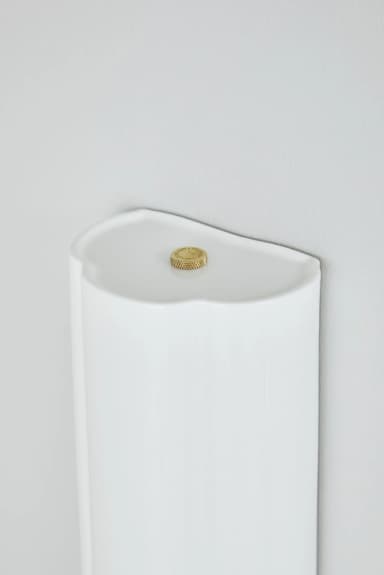 A vertical, cylindrical wall-mounted light fixture featuring a prominent fluted or ribbed design on one side. The fixture is finished in a uniform matte white, suggesting a minimalist and contemporary aesthetic. It is designed to provide indirect illumination, likely casting light upwards or downwards along a wall, depending on its orientation. — 3D model