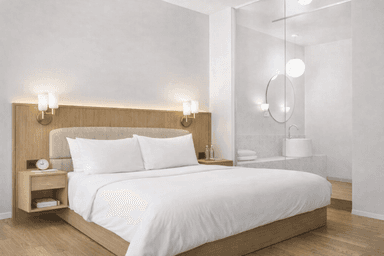 A modern hotel-style bed with a light wooden headboard and frame. The bed features two white pillows and white bedding. Two wall-mounted bedside lamps with golden bases and frosted glass shades are attached to the headboard. Two matching bedside tables are integrated into the headboard structure, each containing a drawer and an open shelf. The bedside table on the left displays a clock and a book, while the one on the right holds two bottles of water. — 3D model