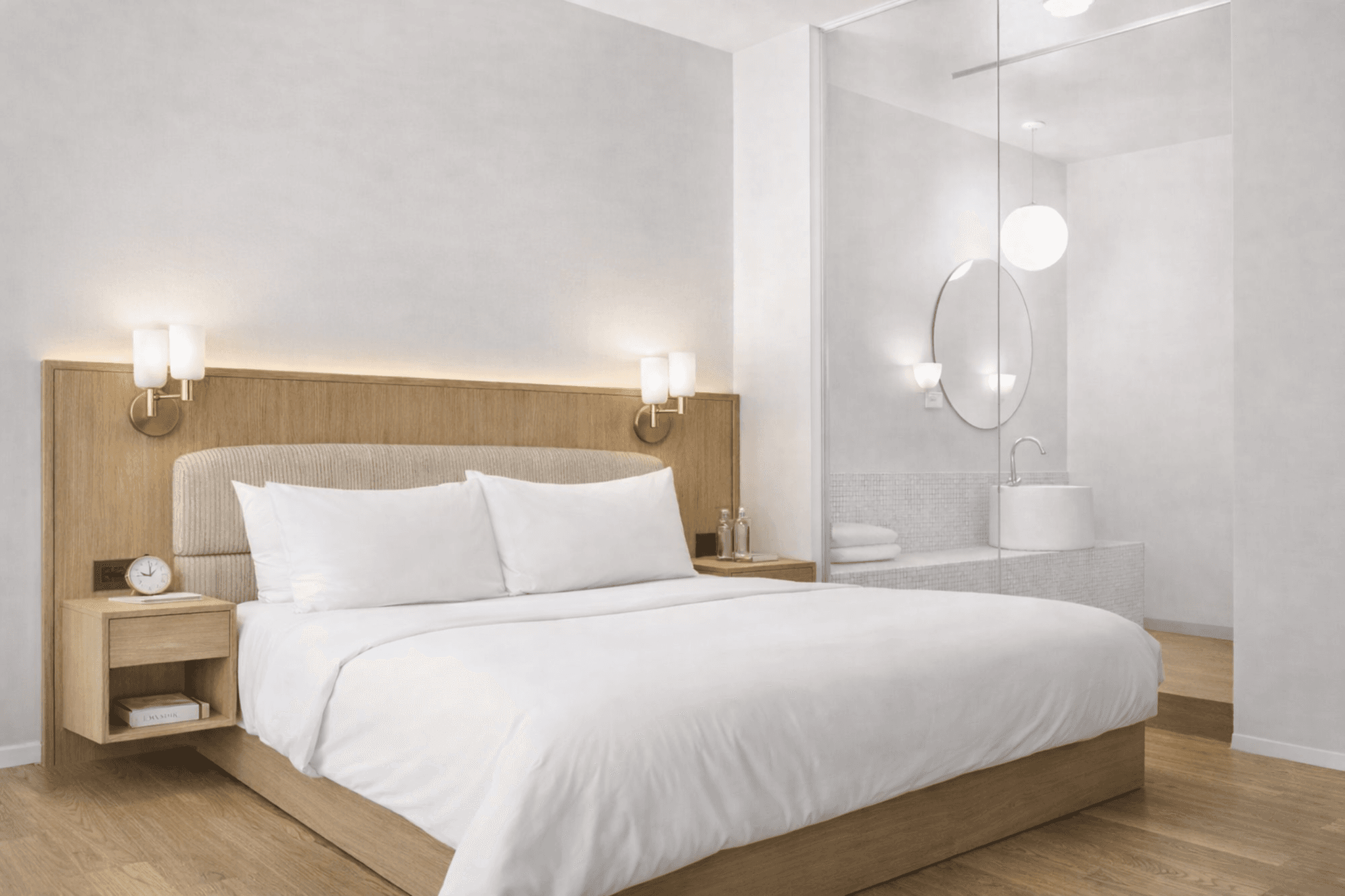 A modern hotel-style bed with a light wooden headboard and frame. The bed features two white pillows and white bedding. Two wall-mounted bedside lamps with golden bases and frosted glass shades are attached to the headboard. Two matching bedside tables are integrated into the headboard structure, each containing a drawer and an open shelf. The bedside table on the left displays a clock and a book, while the one on the right holds two bottles of water. — 3D model