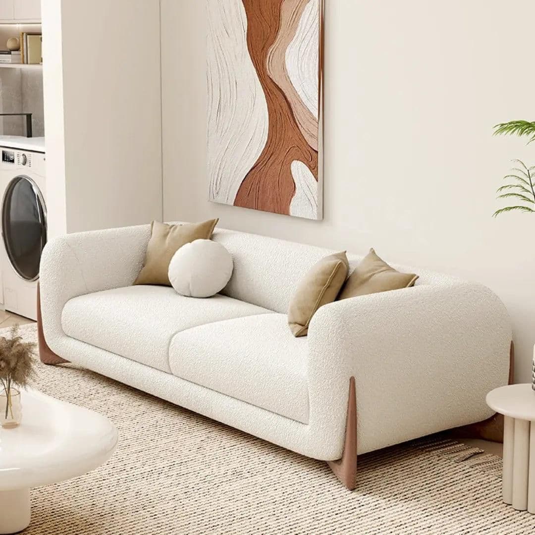 A contemporary two-seater sofa featuring a clean, rounded silhouette and a distinctive integrated wooden base. The sofa is upholstered in a textured, cream-colored fabric, likely bouclé or a similar woven material. It includes two back cushions and three accent pillows—two square and one round—in a complementary light brown hue. The dark wooden base wraps around the lower part of the sofa, providing both structure and a stylish visual contrast to the light upholstery. The design emphasizes comfort with its soft edges and plush appearance. — 3D model