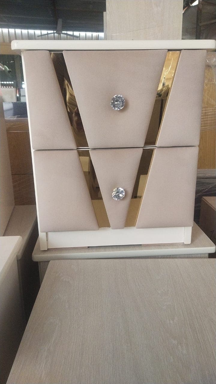 A contemporary nightstand featuring two drawers with upholstered fronts, accented by gold-colored trim and crystal-like knobs. The main body is a light, neutral color, complementing the beige fabric drawers. — 3D model