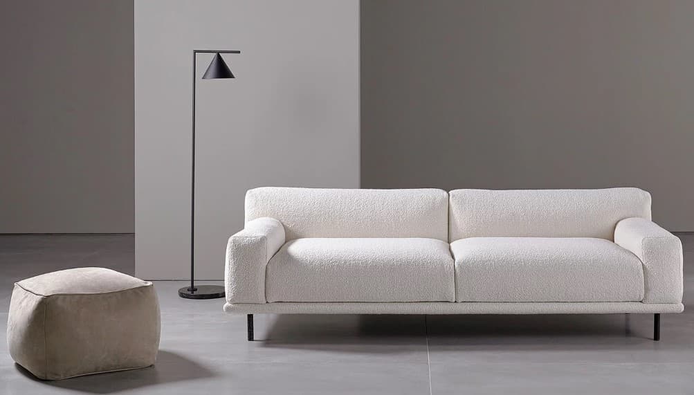 A modern, upholstered sofa with a textured white fabric and a low profile. It features two seat cushions and two back cushions, with gently rounded armrests. The sofa is supported by thin, dark legs. Adjacent to the sofa is a matching upholstered ottoman with a rounded, square shape. — 3D model