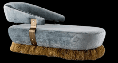 A modern chaise lounge with a curved backrest and an extended seat. The base is embellished with a thick fringe, and the body is accented with a brass-colored strap secured by rivets. The upholstery appears to be a soft, textured fabric. — 3D model