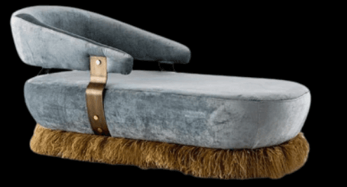A modern chaise lounge with a curved backrest and an extended seat. The base is embellished with a thick fringe, and the body is accented with a brass-colored strap secured by rivets. The upholstery appears to be a soft, textured fabric. — 3D model