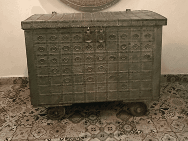 A large, rectangular storage chest with an arched lid, crafted from distressed metal. The entire surface is adorned with an embossed geometric pattern featuring squares with circular motifs. It includes side handles, front latch mechanisms, and small caster wheels for mobility. The finish is a muted dark grey-green, giving it an antique, rustic appearance. — 3D model