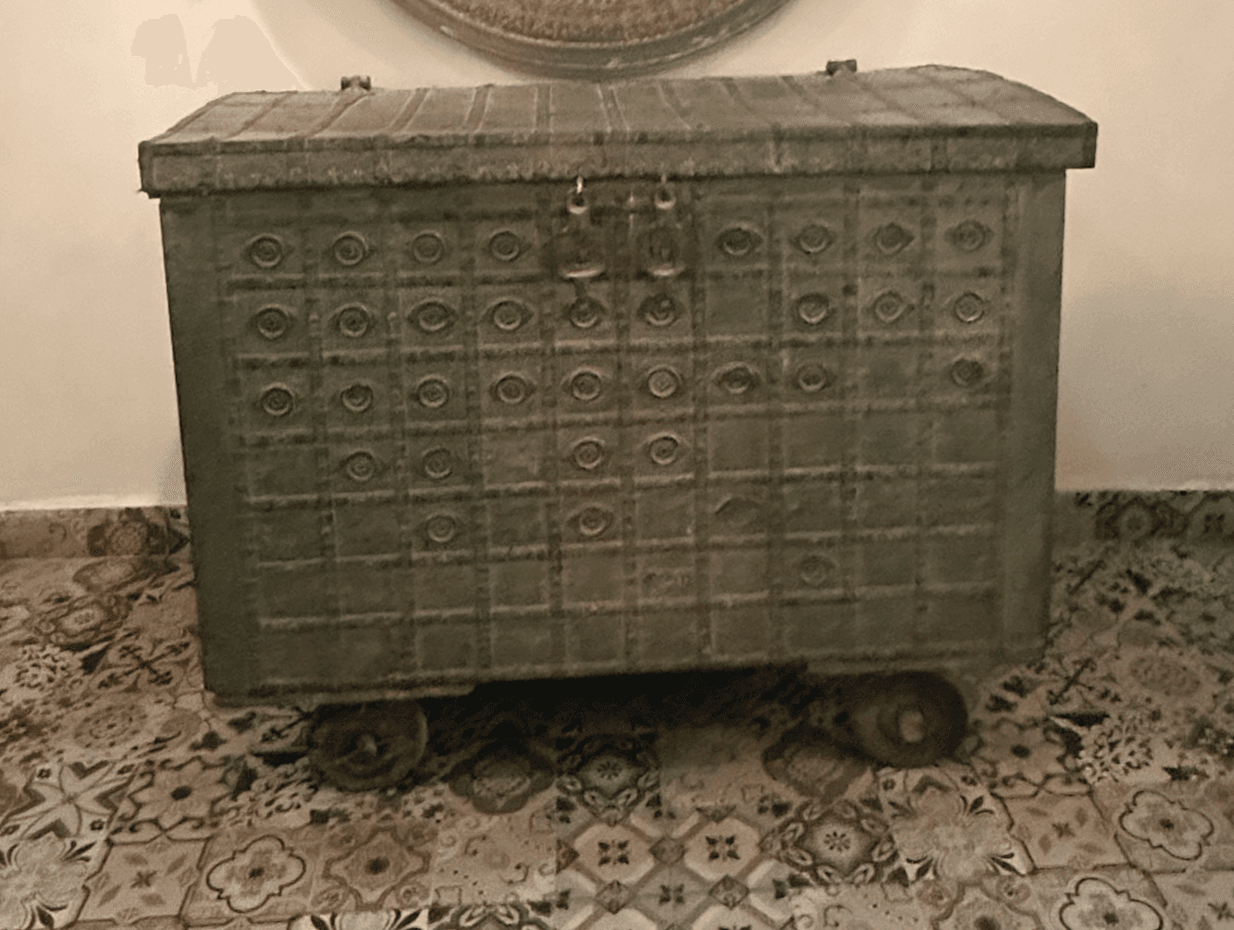 A large, rectangular storage chest with an arched lid, crafted from distressed metal. The entire surface is adorned with an embossed geometric pattern featuring squares with circular motifs. It includes side handles, front latch mechanisms, and small caster wheels for mobility. The finish is a muted dark grey-green, giving it an antique, rustic appearance. — 3D model