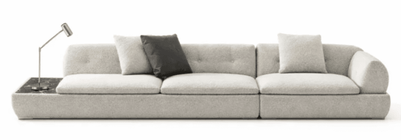 A long, contemporary modular sofa upholstered in a light, textured fabric, likely bouclé. It features multiple seat and back cushions, some with button tufting. An integrated dark, veined marble side table is positioned at one end, supporting a small, silver-toned task lamp. The sofa has a low-profile design with soft, rounded edges. — 3D model