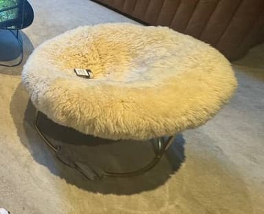 A contemporary circular ottoman featuring a plush, cream-colored faux fur cushion and a minimalist gold-finished metal base. The cushion has a distinctive donut-like shape with a central indentation, providing a soft and inviting surface. The base consists of two intersecting curved metal bars, creating a stable yet open and airy design. — 3D model