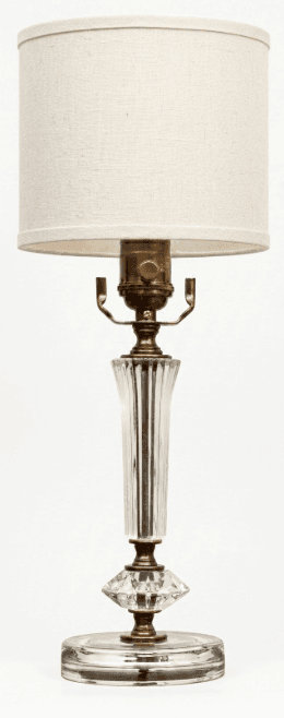 A traditional table lamp featuring a clear glass body with fluted and faceted details, set on a round glass base. It is accented with brass-toned metal components, including the harp and socket. The lamp is topped with a simple, cylindrical off-white fabric drum shade. — 3D model