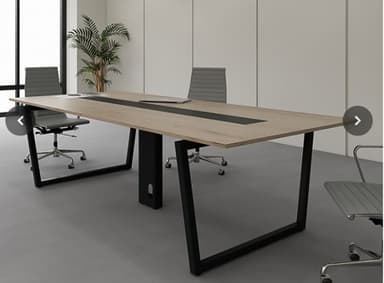 A long, rectangular conference table featuring a light wood-grain laminate top with a prominent black central channel, likely for power and data. The table is supported by two black, U-shaped metal legs at the ends and a central black modesty panel or support column, also with a cutout for cable management. Its design is clean and contemporary. — 3D model
