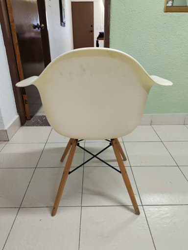 An off-white, molded plastic armchair with integrated armrests and a 'Eiffel Tower' style base consisting of splayed wooden dowel legs reinforced with black metal cross-supports. The chair is positioned on a light-colored tiled floor. — 3D model