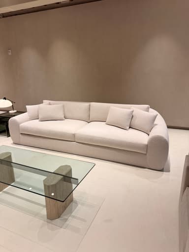 A long, contemporary upholstered sofa featuring a light cream fabric, soft rounded armrests, and a low-profile back, complete with multiple seat, back, and throw cushions. — 3D model