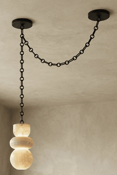 A contemporary pendant light featuring a sculptural shade crafted from translucent alabaster, suspended by a black metal chain system. The shade is composed of three stacked, organic, rounded forms, illuminated from within to highlight the natural veining and luminous quality of the stone. The suspension system consists of a long chain dropping from one ceiling mount, connecting to the light fixture, and a shorter chain extending horizontally to a second ceiling mount, creating a distinctive draped effect. — 3D model