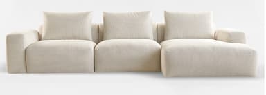 A contemporary, plush, light beige sectional sofa featuring a modular design with clean lines and a low profile. It consists of multiple sections, including a three-seat segment and an extended chaise lounge, all upholstered in a textured fabric. The backrests are composed of loose, rectangular cushions, complementing the soft, rounded appearance of the base and arms. — 3D model