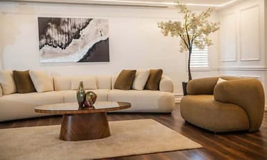 A modular, curved sectional sofa upholstered in a light beige fabric. It features a low profile, rounded arms, and a series of connecting sections that create a flowing silhouette. The sofa is adorned with several throw pillows in complementary shades of cream and brown. — 3D model