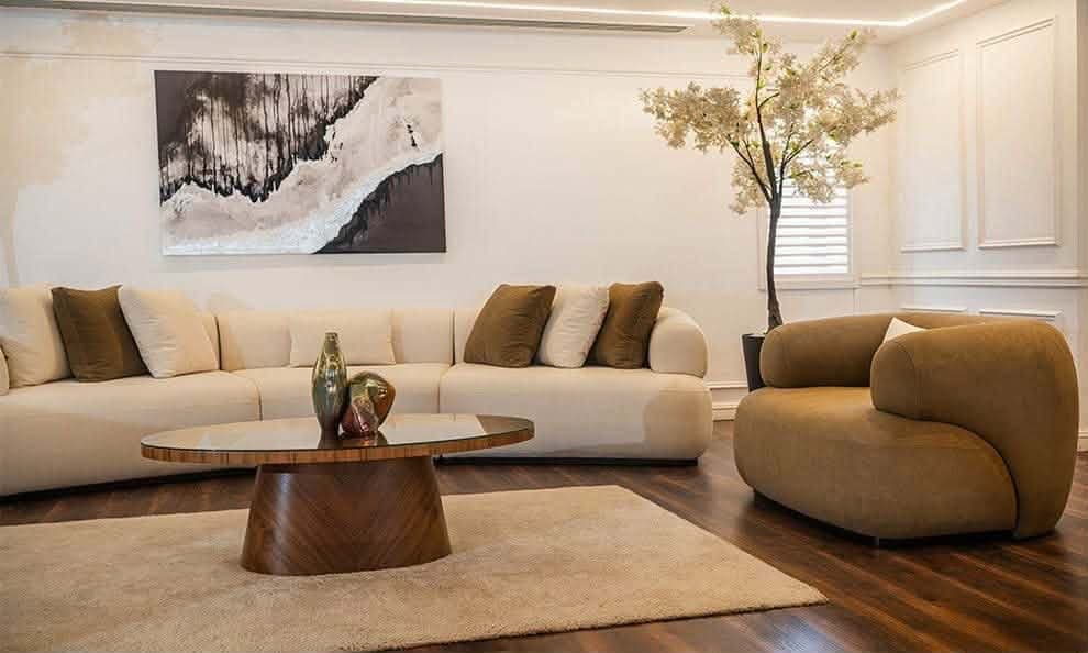 A modular, curved sectional sofa upholstered in a light beige fabric. It features a low profile, rounded arms, and a series of connecting sections that create a flowing silhouette. The sofa is adorned with several throw pillows in complementary shades of cream and brown. — 3D model