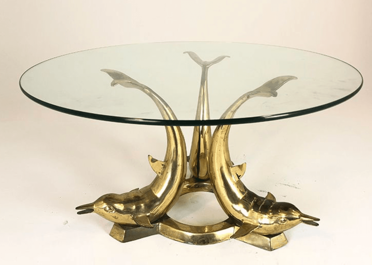 A round glass-top coffee table with a distinctive base. The base is constructed from polished brass and features three stylized dolphin figures. Each dolphin is depicted in a dynamic pose, supporting the glass tabletop with its tail or fins. The dolphins' bodies curve upwards, forming the central support structure of the table, connected by a circular brass element at their bases. — 3D model