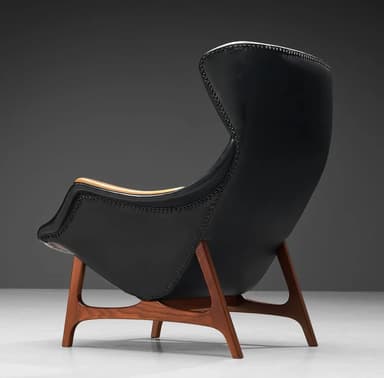 A high-backed armchair with a distinctive, sculptural form. The seat and backrest are upholstered in black material, possibly leather, and accented with numerous small decorative studs along the edges. The arms appear to be crafted from a lighter-colored wood, contrasting with the main upholstery. The chair is supported by a sculpted wooden base with a dark wood finish, featuring angled legs that provide stability. — 3D model