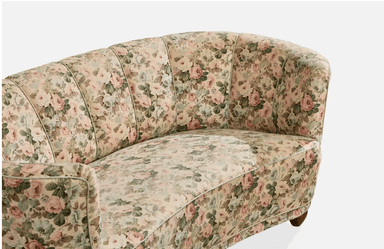 A vintage-style, curved sofa upholstered in a floral fabric with a pastel color palette. The sofa features a rounded backrest with vertical channeling and integrated armrests that curve inwards. It is supported by four short, tapered wooden legs. — 3D model