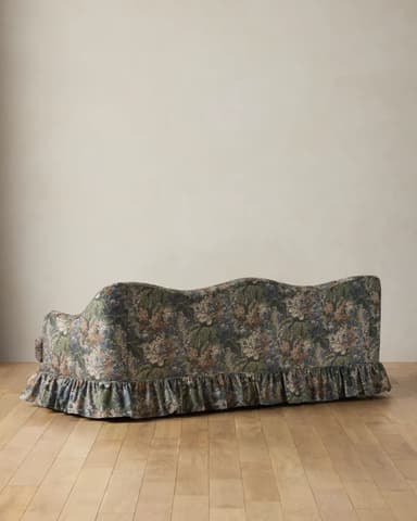 A three-seater sofa featuring a distinctive scalloped backrest and a fully upholstered frame. It is covered in a dark floral fabric with prominent green leaves, blue and white flowers, and hints of orange. The sofa has rolled arms and a gathered, ruffled skirt concealing its legs. — 3D model