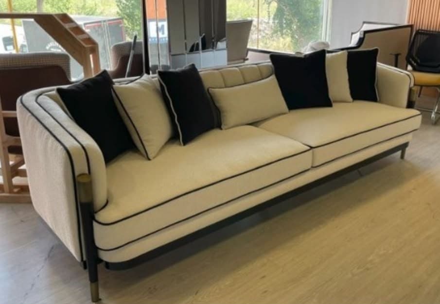 A contemporary three-seater sofa featuring a cream or off-white upholstered body with distinctive black piping along all edges. The sofa has a structured, rounded armrest design and a backrest with subtle channel tufting. It stands on slim, straight black metal legs with brass-finished caps, giving it a light, elevated appearance. The sofa is accompanied by a mix of black and cream throw pillows, also accented with black piping. — 3D model