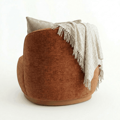 A contemporary armchair with a rounded silhouette, upholstered in a textured, rust-colored fabric. It features a low profile with a visible wooden base. A textured throw blanket with fringes and a decorative pillow are draped over the back and armrest. — 3D model
