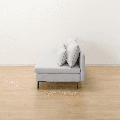 A contemporary armless chaise loveseat, upholstered in a light gray textured fabric. It features a low-profile design with a deep, elongated seat on the right side, resembling a chaise. The seating is supported by minimalist black metal legs, and the piece includes two large back cushions and two smaller rectangular throw pillows, all with subtle piped edges. — 3D model