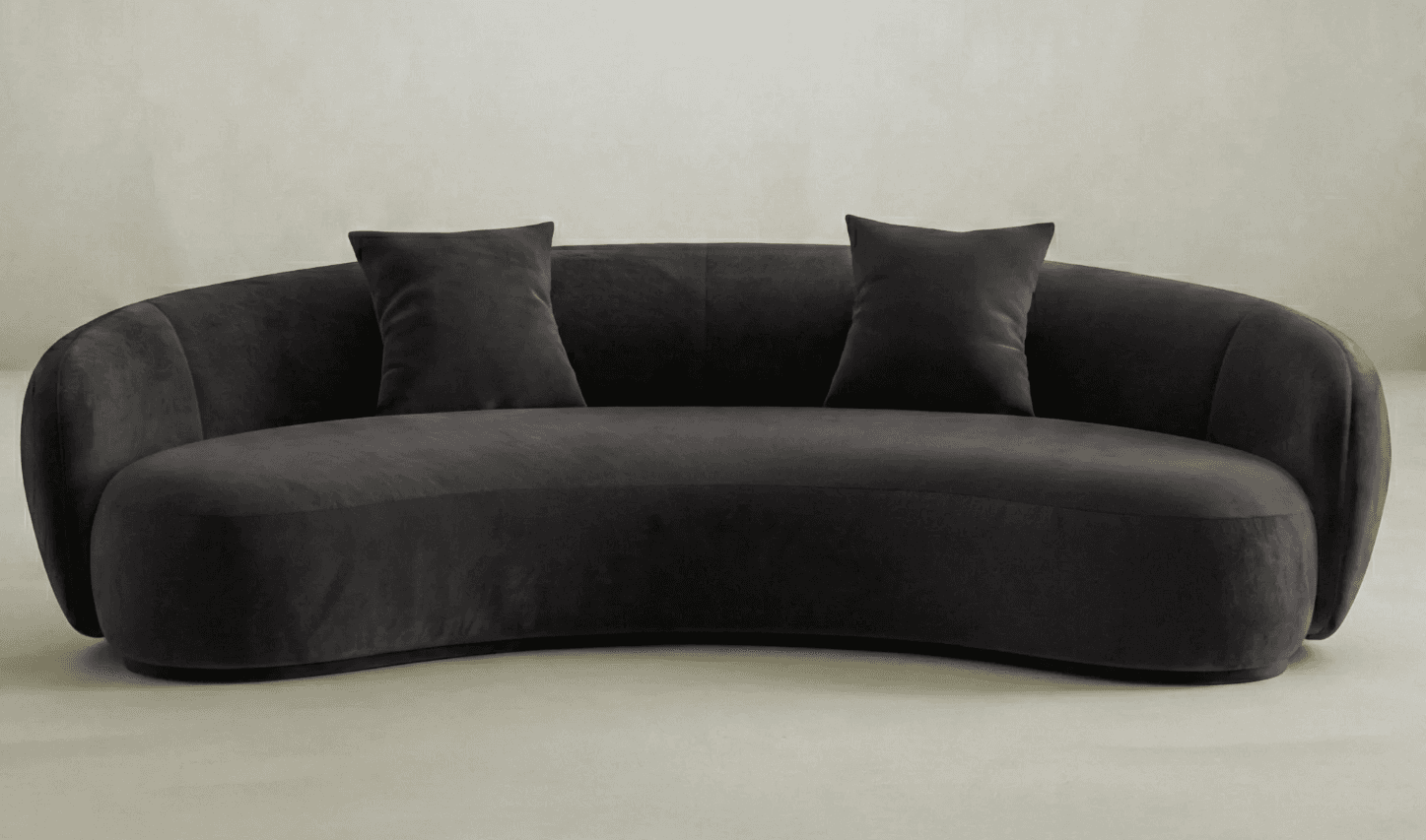A curved, modern sofa upholstered in a dark grey, plush fabric. The sofa features a continuous, rounded backrest that extends into armrests on either side. The seat is also curved to follow the overall shape. Two matching square throw pillows are placed on the seat. The design is minimalist with subtle vertical tufting on the backrest. — 3D model