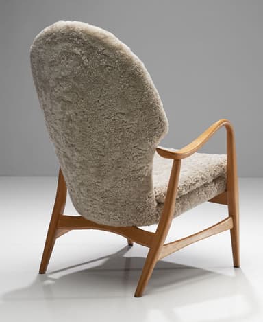 A high-backed armchair with a wooden frame and plush sheepskin upholstery. The frame consists of curved wooden arms and legs. The seat and back are fully upholstered in a textured, cream-colored sheepskin material, giving it a soft and inviting appearance. The design suggests a focus on comfort and organic forms. — 3D model