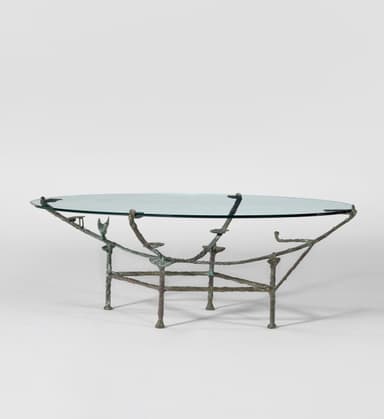 An oval coffee table with a clear glass top supported by a sculptural metal base. The base appears to be cast bronze or a similar metal, with an organic, somewhat branching structure that mimics natural forms. It features four legs that connect to a lower frame, supporting the tabletop via four points. — 3D model