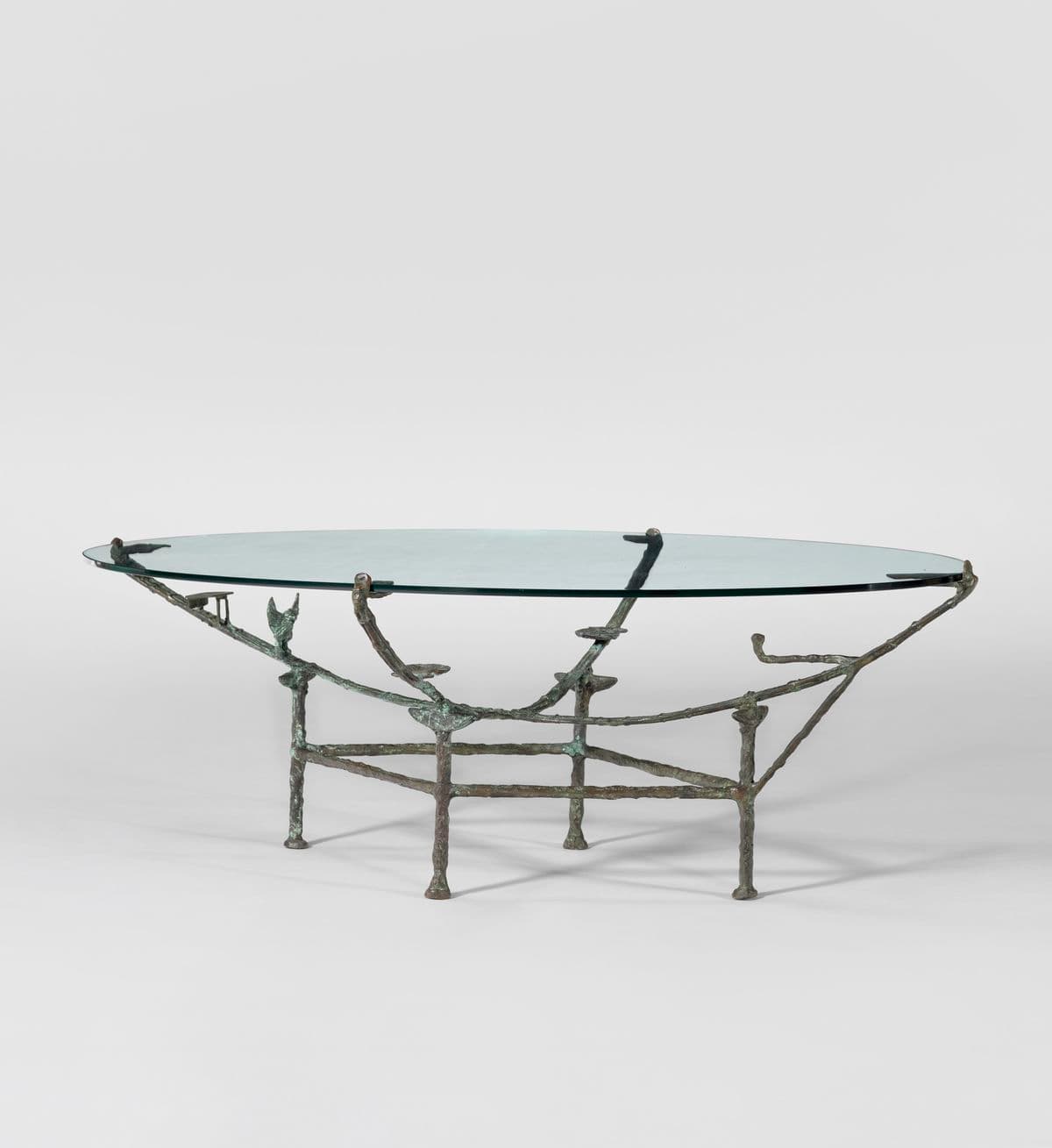 An oval coffee table with a clear glass top supported by a sculptural metal base. The base appears to be cast bronze or a similar metal, with an organic, somewhat branching structure that mimics natural forms. It features four legs that connect to a lower frame, supporting the tabletop via four points. — 3D model