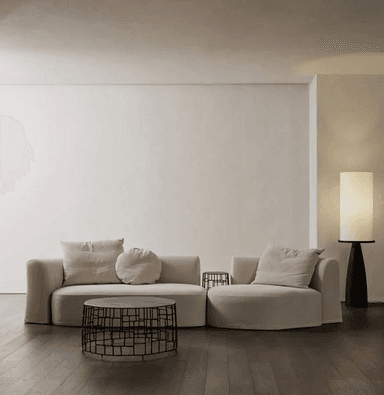 A modular sectional sofa with a curved chaise lounge on the right and a straight section on the left. It features a low profile, rounded arms and base, and is upholstered in a light beige fabric. The sofa is accompanied by three decorative pillows: one square in the same fabric as the sofa, one rectangular, and one round, both also in a similar light beige fabric. — 3D model