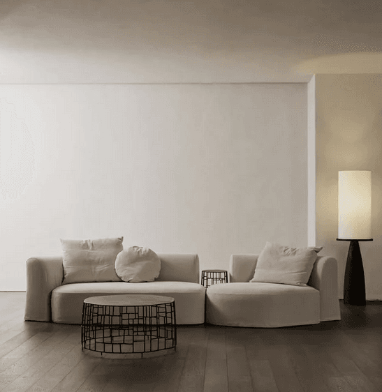A modular sectional sofa with a curved chaise lounge on the right and a straight section on the left. It features a low profile, rounded arms and base, and is upholstered in a light beige fabric. The sofa is accompanied by three decorative pillows: one square in the same fabric as the sofa, one rectangular, and one round, both also in a similar light beige fabric. — 3D model