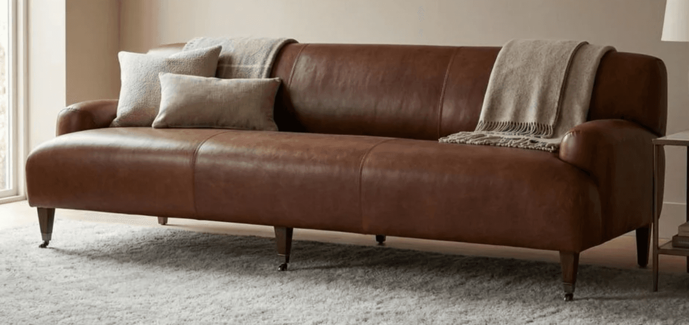 A traditional brown leather sofa featuring classic rolled arms, a single back cushion with vertical stitching, and three integrated seat cushions. The sofa stands on tapered dark wooden legs finished with brass casters. — 3D model