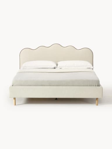A cream-colored upholstered bed frame with a distinctive scalloped headboard. The entire frame, including the headboard, side rails, and footboard, is covered in a soft, textured fabric. The headboard features a wavy top edge with contrasting piping. The bed stands on four slender, light-toned tapered wooden legs. A white mattress and a light gray throw are visible on the bed. — 3D model